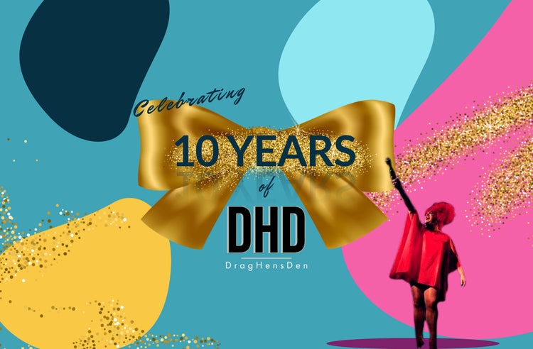 Sheila Fitzpatrick, one of the hosts of the show, is seen pointing to a banner that reads "Celebrating 10 years of DragHensDen". Sheila is standing on a stage with a microphone in her hand, dressed in a long-sleeved red dress and red headpiece. The background shows bright colours and glitter. The image captures Sheila acknowledging the milestone of the show's 10 year anniversary.